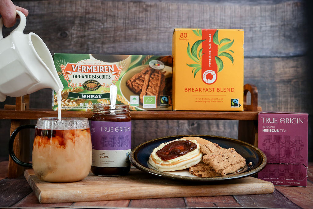 Person pouring Traidcraft Tea into a mug with Fairtrade biscuits and a scone with fair trade guava jam on a wooden surface.