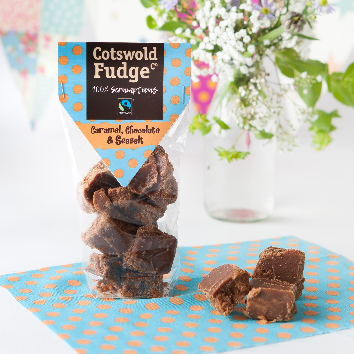 A bag of 100% scrumptious Fairtrade Caramel, Chocolate and Seasalt Fudge made by Cotswold Fudge Co. The bag is transparent with chunks of fudge on display. The bag is sitting on a light blue napkin with orange polkadots with some flowers in the background.