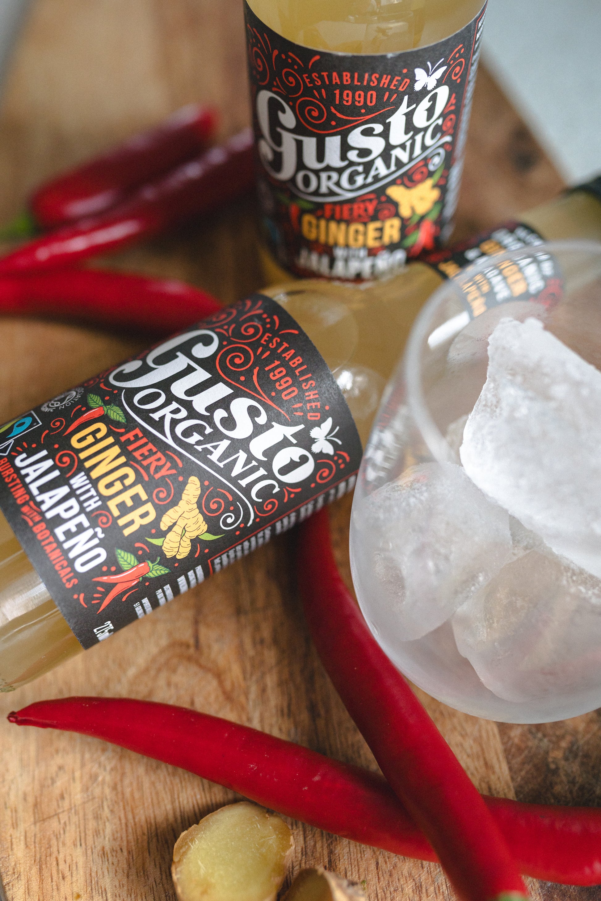 Two bottles of Fairtrade Gusto Organic Ginger with Jalapeno soft drink on a wooden surface surrounded with chillies and a glass with ice.