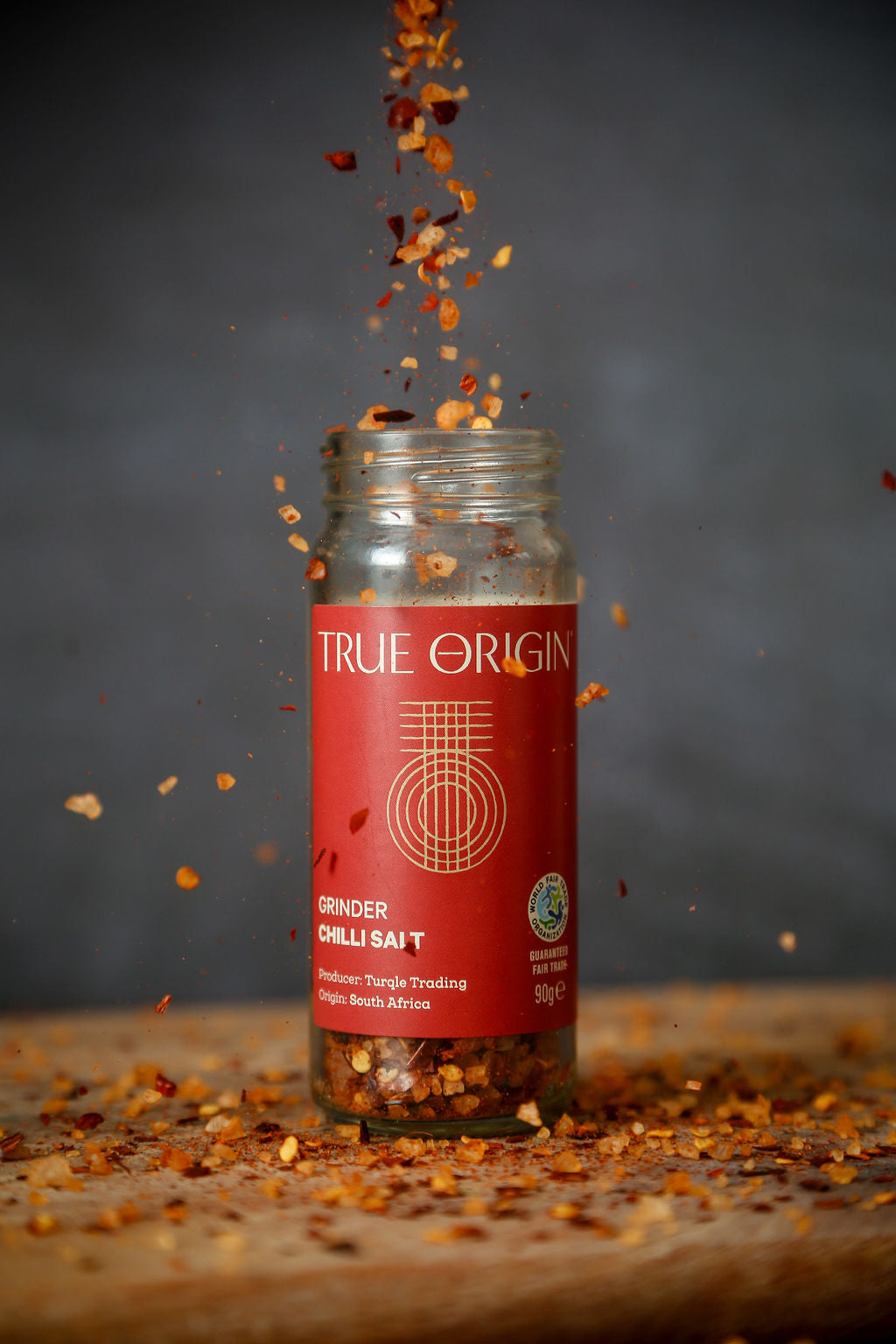 Open jar of True Origin chilli salt on a wooden chopping board with chilli flakes and salt crystals being poured into it from above