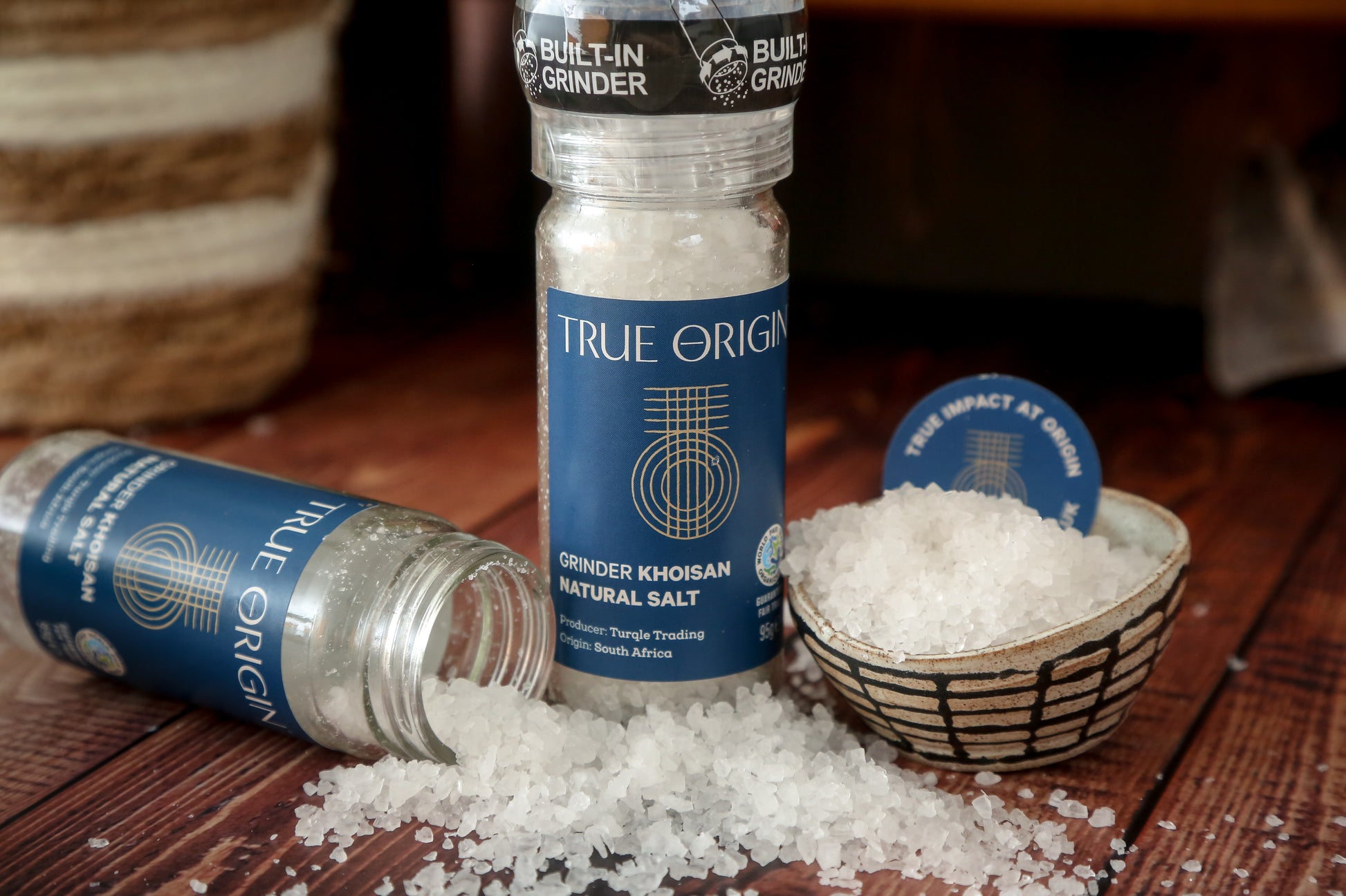 A clear glass bottle with a built-in grinder containing natural Khoisan salt, labelled 'True Origin'.  There is an opened jar of salt crystals being poured in a bowl and on the worktop.