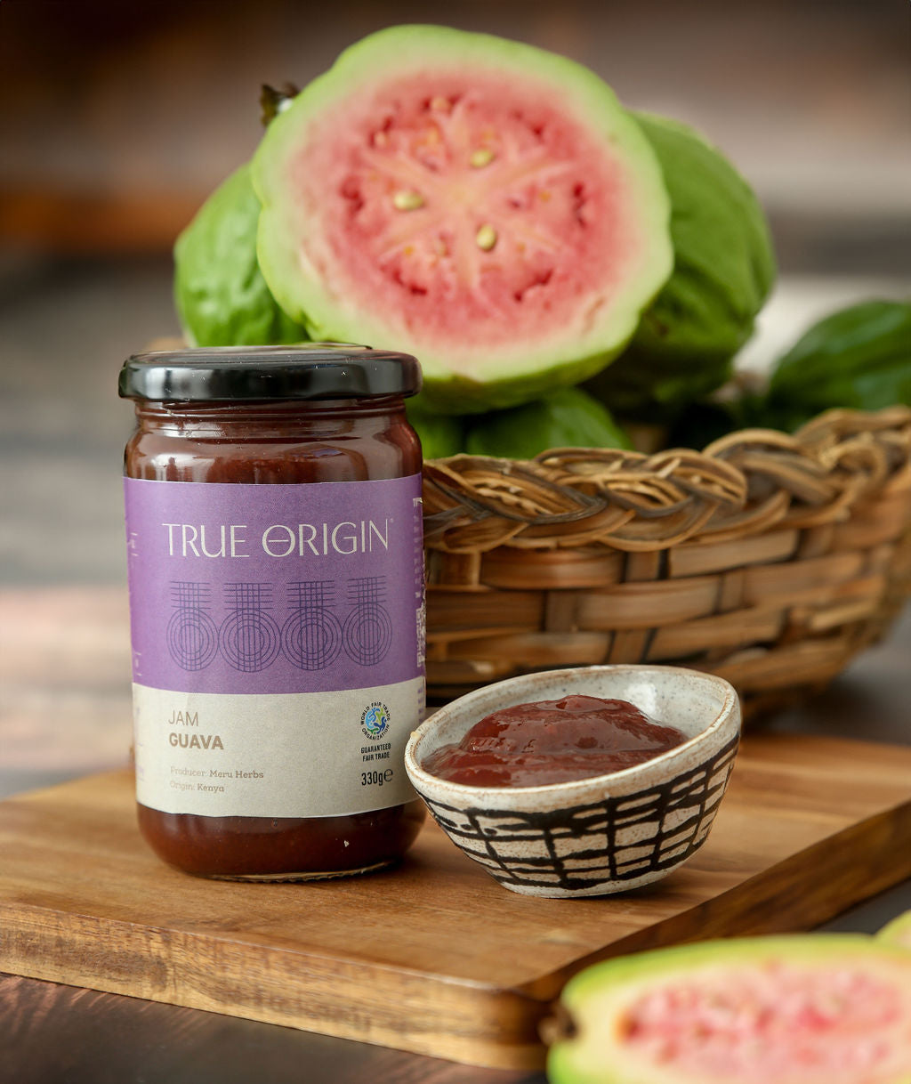 A jar of guava jam with a label that reads 'TRUE ORIGIN' and 'JAM GUAVA 330G'. It is sitting on a wooden surface with a bowl of guava jam and a bowl of fresh and cut guavas.