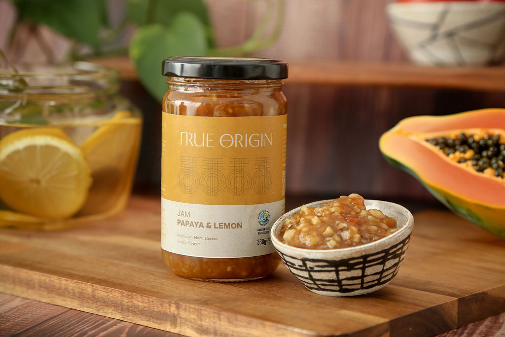 A jar of True Origin papaya and lemon jam on a wooden chopping board with an open papaya and a bowl with papaya jam in it. There is a also a bowl containing lemon slices.