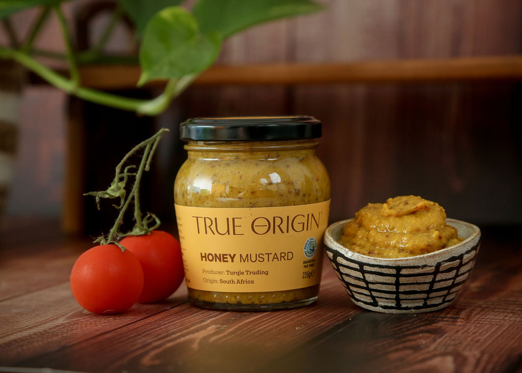 A jar of yellow premium True Origin Honey Mustard beside a bowl full of mustard at one side and two tomatoes on a vine at the other side on a wooden surface with a plant in the background.