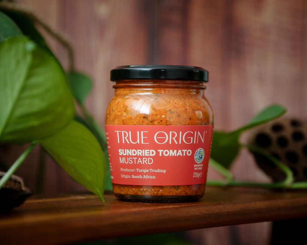 An unopened red jar of True Origin Sundried Tomato Mustard on a wooden shelf with some leaves in the background.