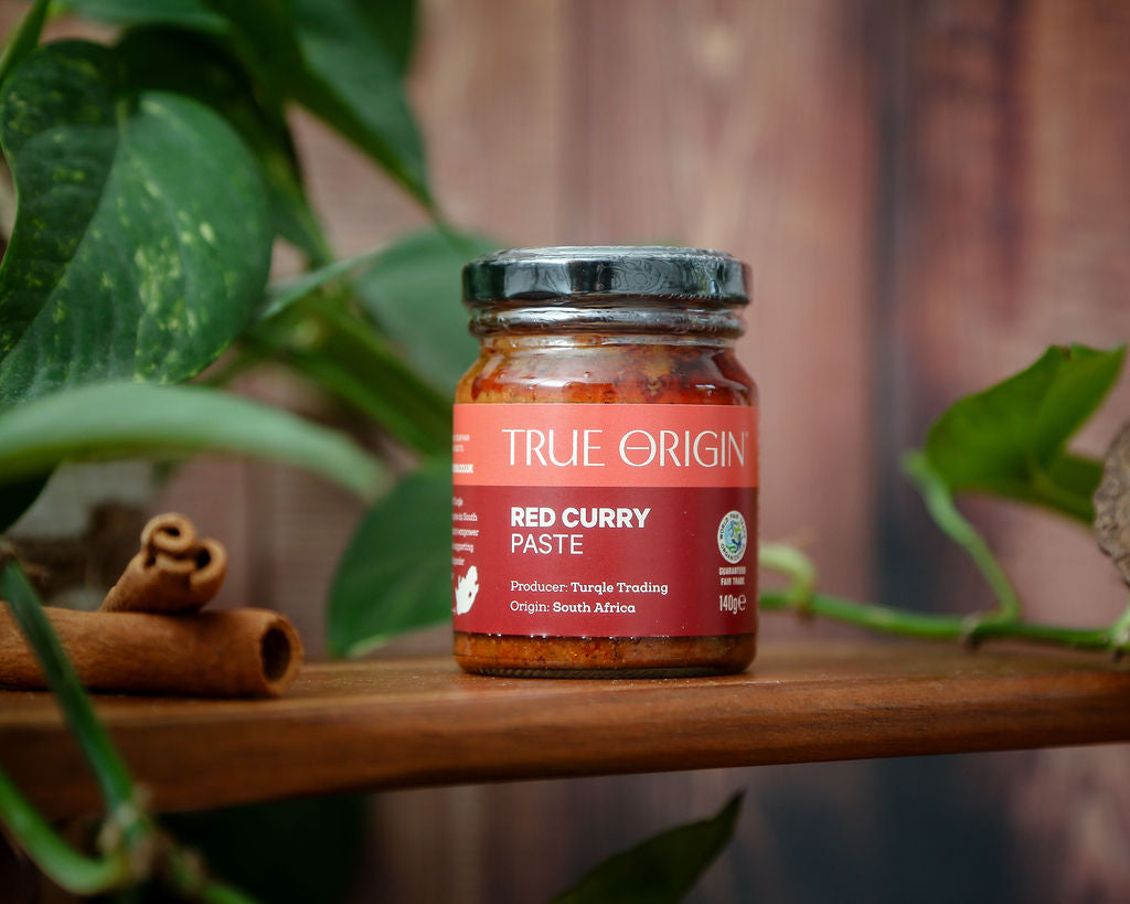An unopened red jar of True Origin Red Curry Paste on a wooden shelf next to two cinnamon sticks and some leaves in the background.
