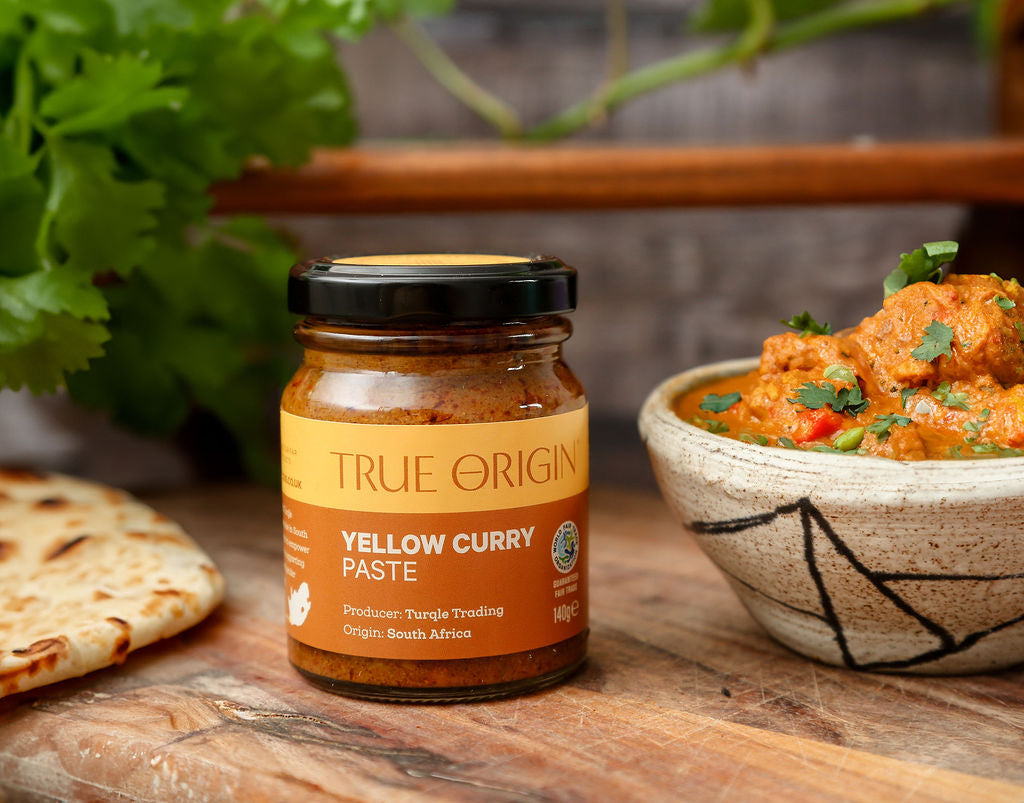 An unopened yellow jar of True Origin Yellow Curry Paste on a wooden surface next to a bowl of yellow curry and a naan bread with some parsley in the background.