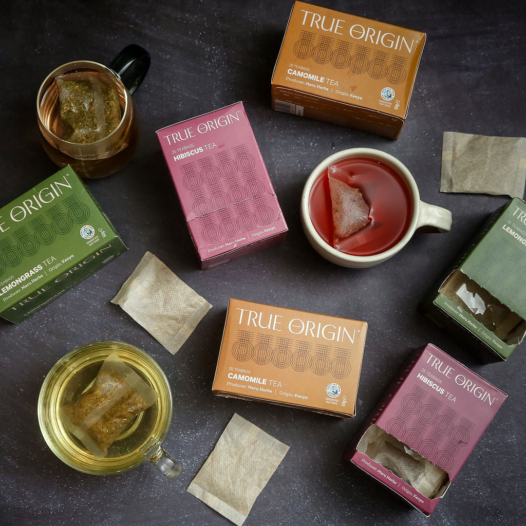 True Origin tea boxes and bags on a dark surface with herbal tea in cups.
