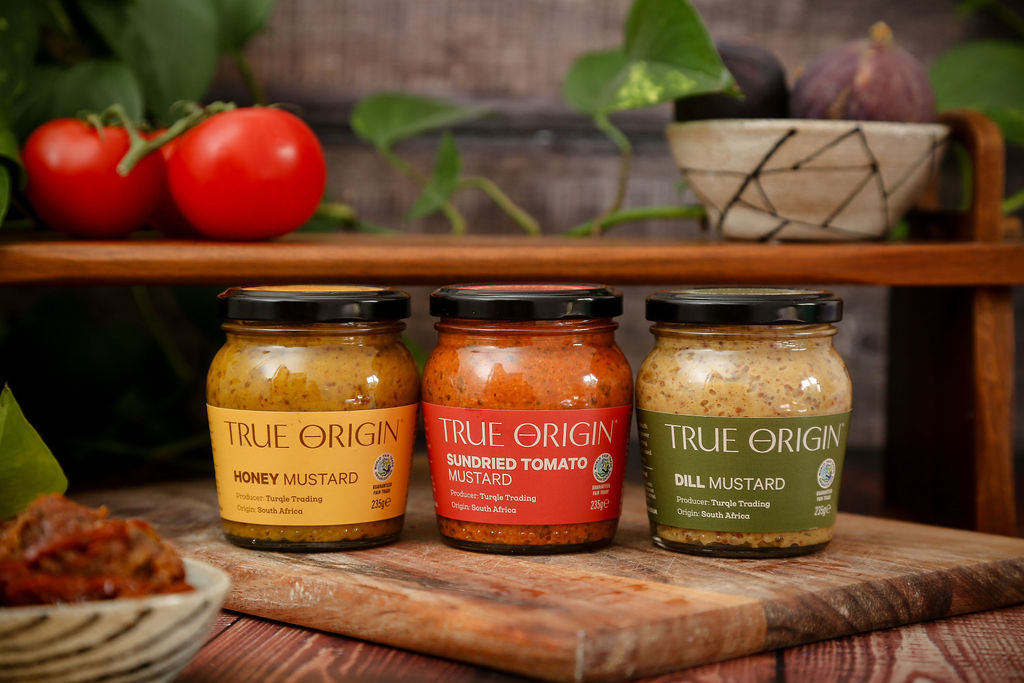 Yellow Honey, Red Sundried Tomato and a Green Dill Mustard jars in a row on a wooden chopping board with tomatoes and figs in a bowl in the background.