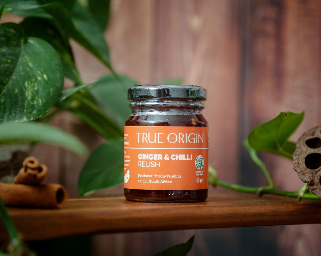 An orange jar of True Origin Ginger and Chilli relish sitting on a wooden shelf next to some cinnamon and some green leaves in the background.