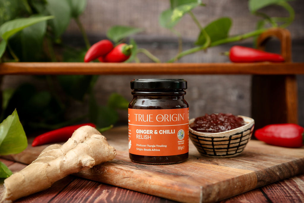 An orange jar of True Origin Ginger and Chilli relish next to some relish in a bowl and some ginger with some chillies in the background.
