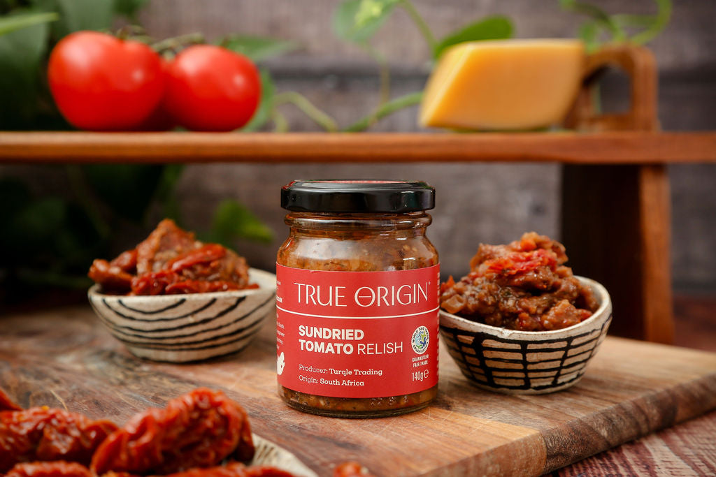 A red jar of True Origin Sundried Tomato relish next to some relish in a bowl and some tomatoes and cheese in the background.