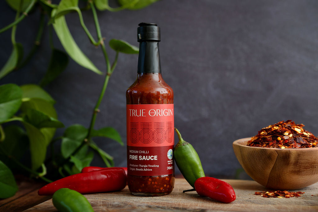 Bottle of True Origin fire sauce with peppers and a bowl of spices on a wooden surface.