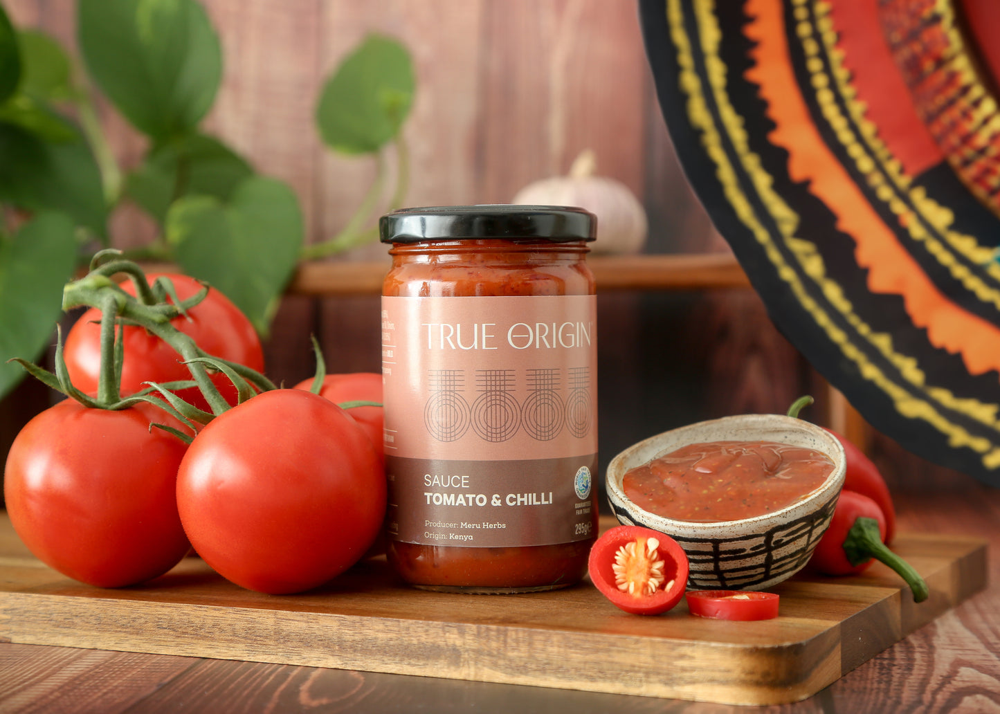 A jar of True Origin brand Tomato & Chilli sauce produced by Meru Herbs in Kenya. The jar is sitting on a wooden chopping board next to some fresh tomatoes and fresh chillies and a bowl containing that tomato and chilli sauce.