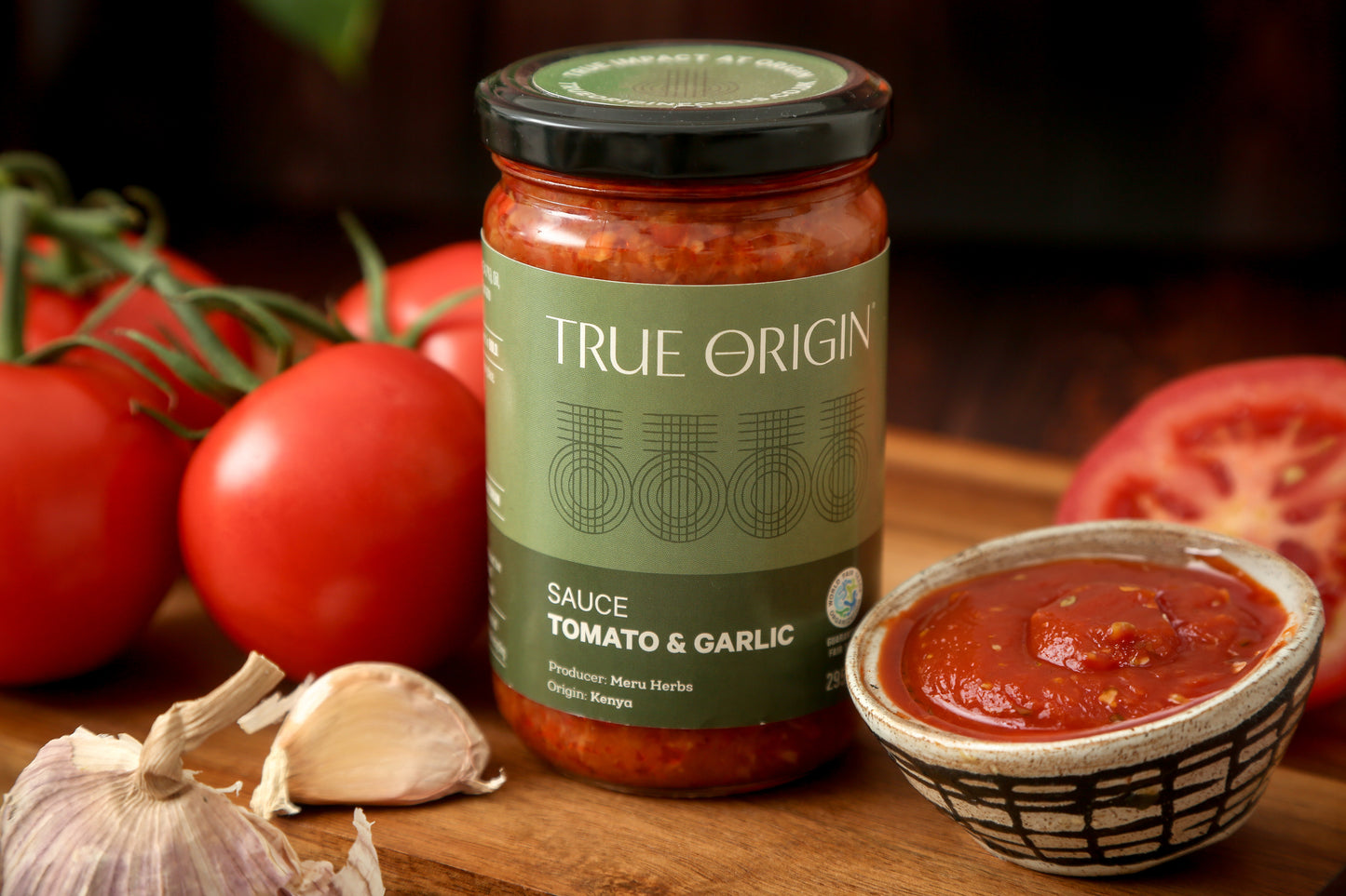 A jar of True Origin Tomato and Garlic sauce sitting on a wooden surface with a bowl of tomato and garlic sauce and some fresh tomatoes and garlic.