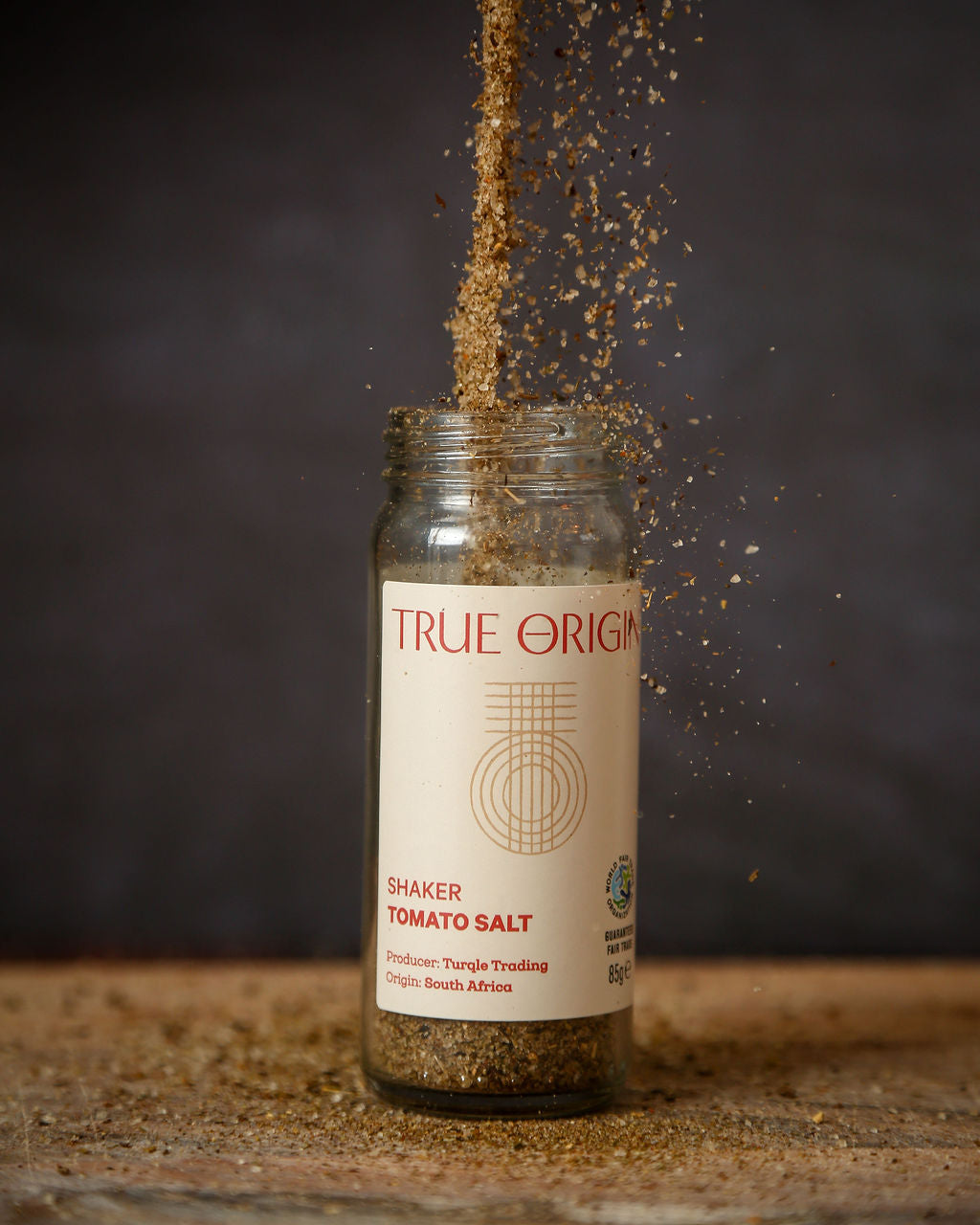 An open white glass jar of tomato salt with fair trade tomato salt being poured into it from above and sprinkling onto a chopping board.