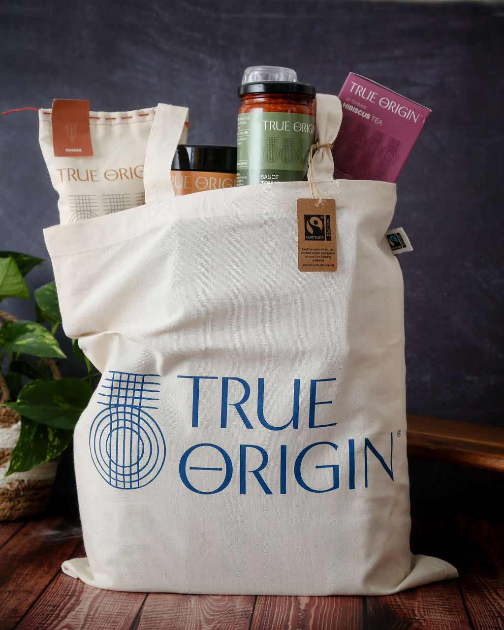 A cream Fairtrade cotton tote bag with the blue stacked True Origin logo with some True Origin products sticking out the top.