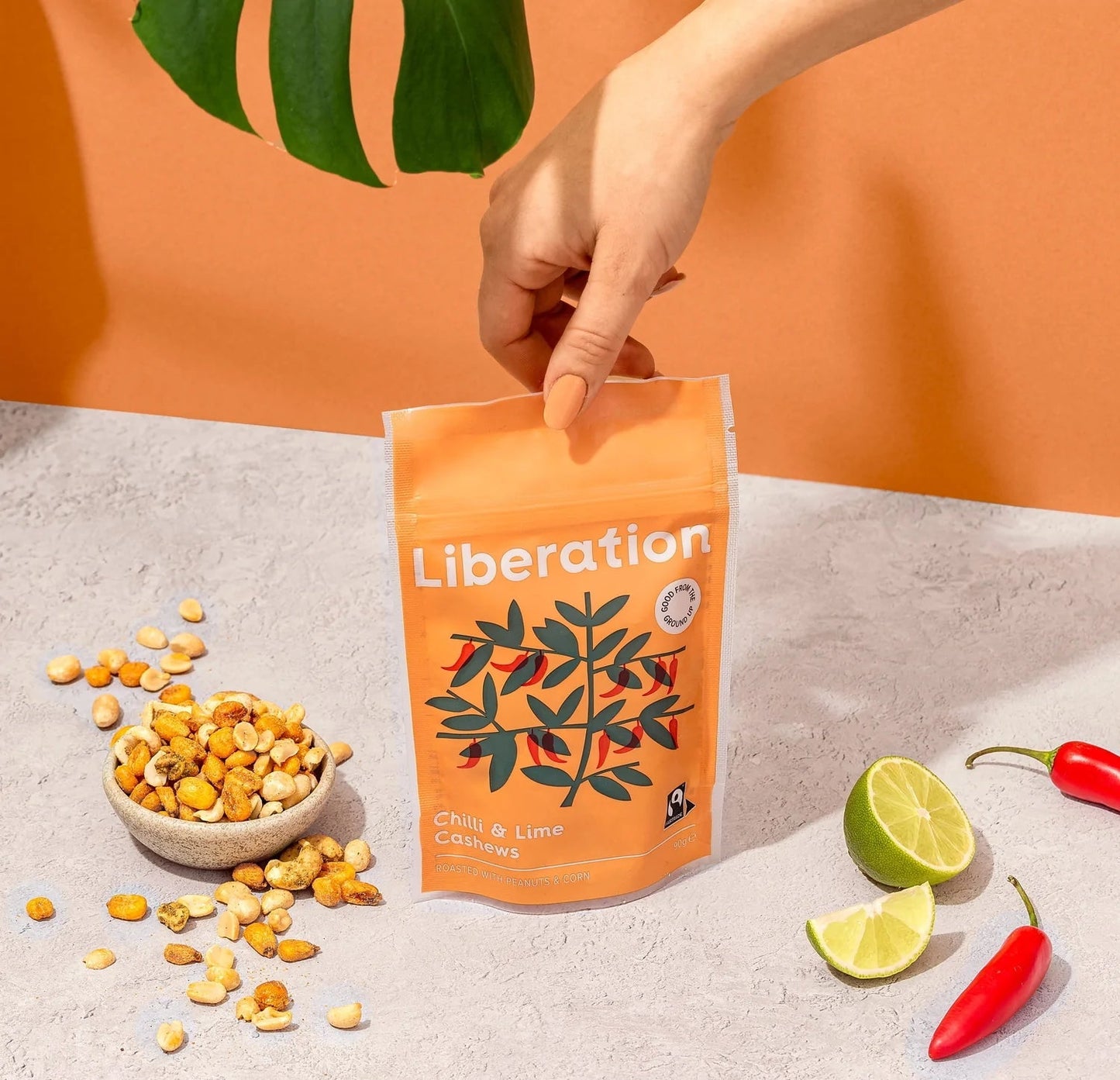 Packaging of Liberation Foods Chilli & Lime Cashews with an illustration of cashew nuts and chilli peppers on an orange background. There are some peanuts, some chillies and a cut fresh lime beside the bag.