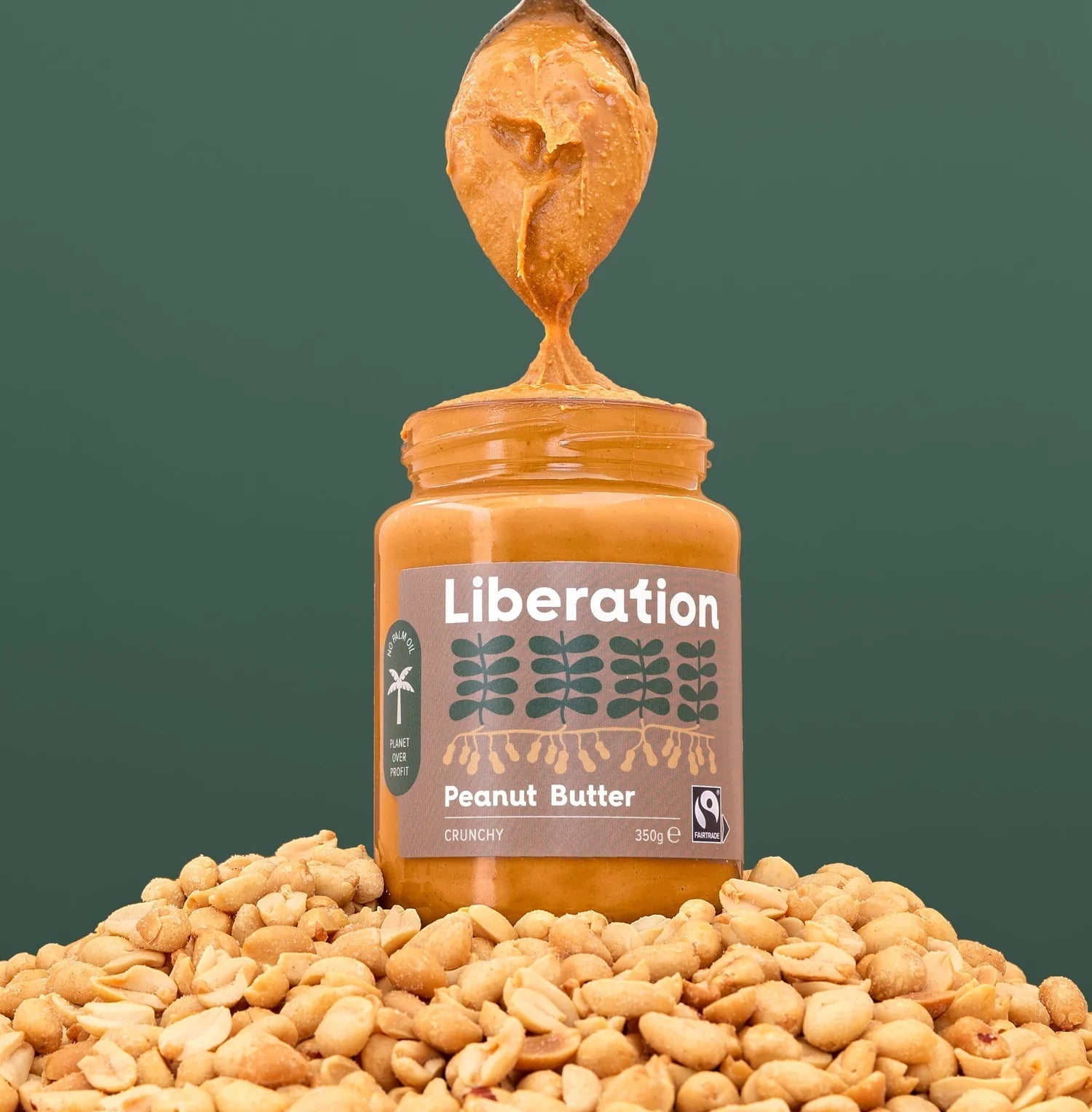 A jar of Liberation Foods Fairtrade Peanut Butter sitting on a pile of peanuts with a spoon taking some peanut butter out of the jar.