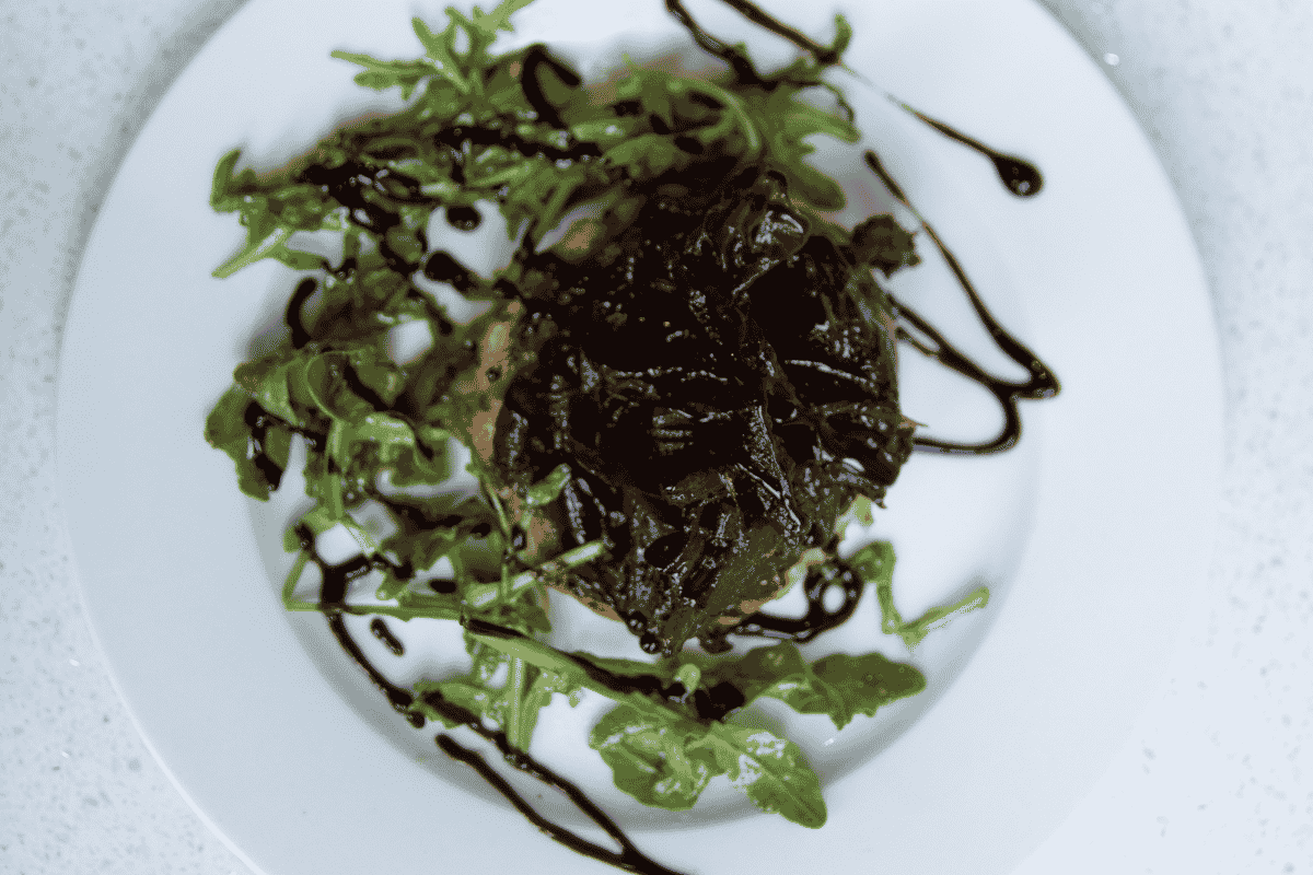 Caramelised onions are dark and glossy on top of a pastry tart on a white plate with green salad leaves and balsamic glaze design  