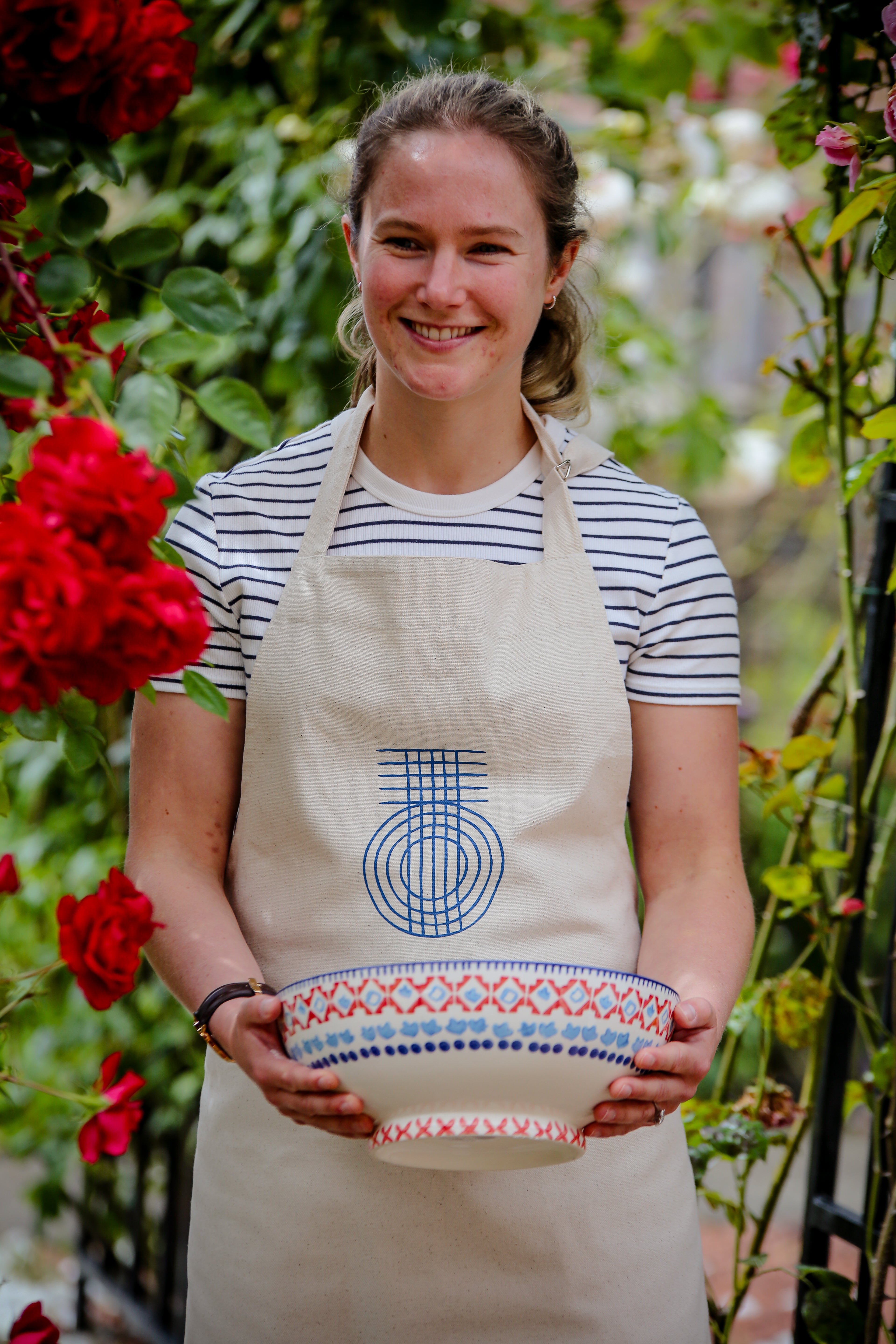 A woman wearing a beige True Origin apron carrying a bowl through a flower garden.