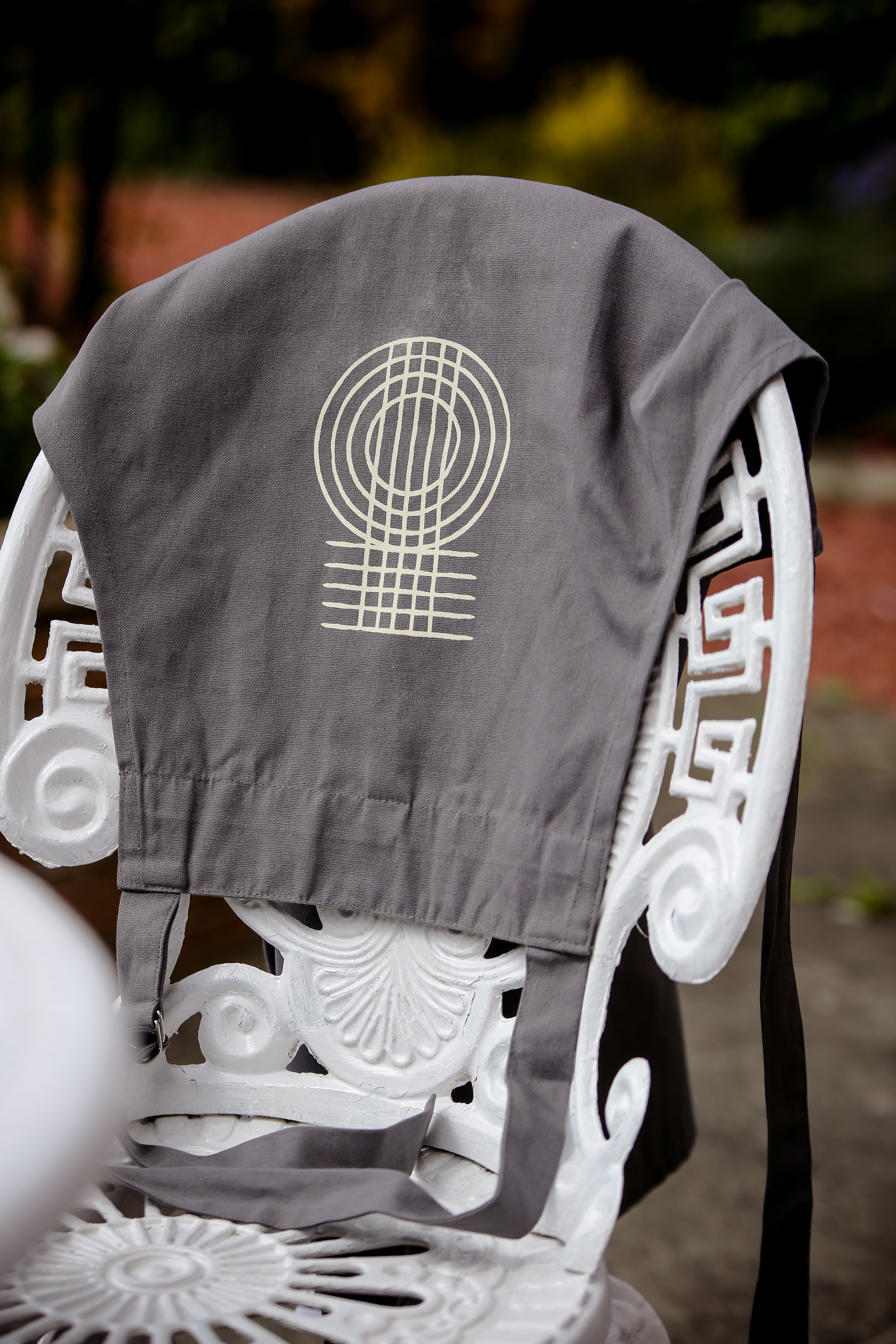 A Fairtrade cotton grey apron over the back of a patio chair with the True Origin logo showing.