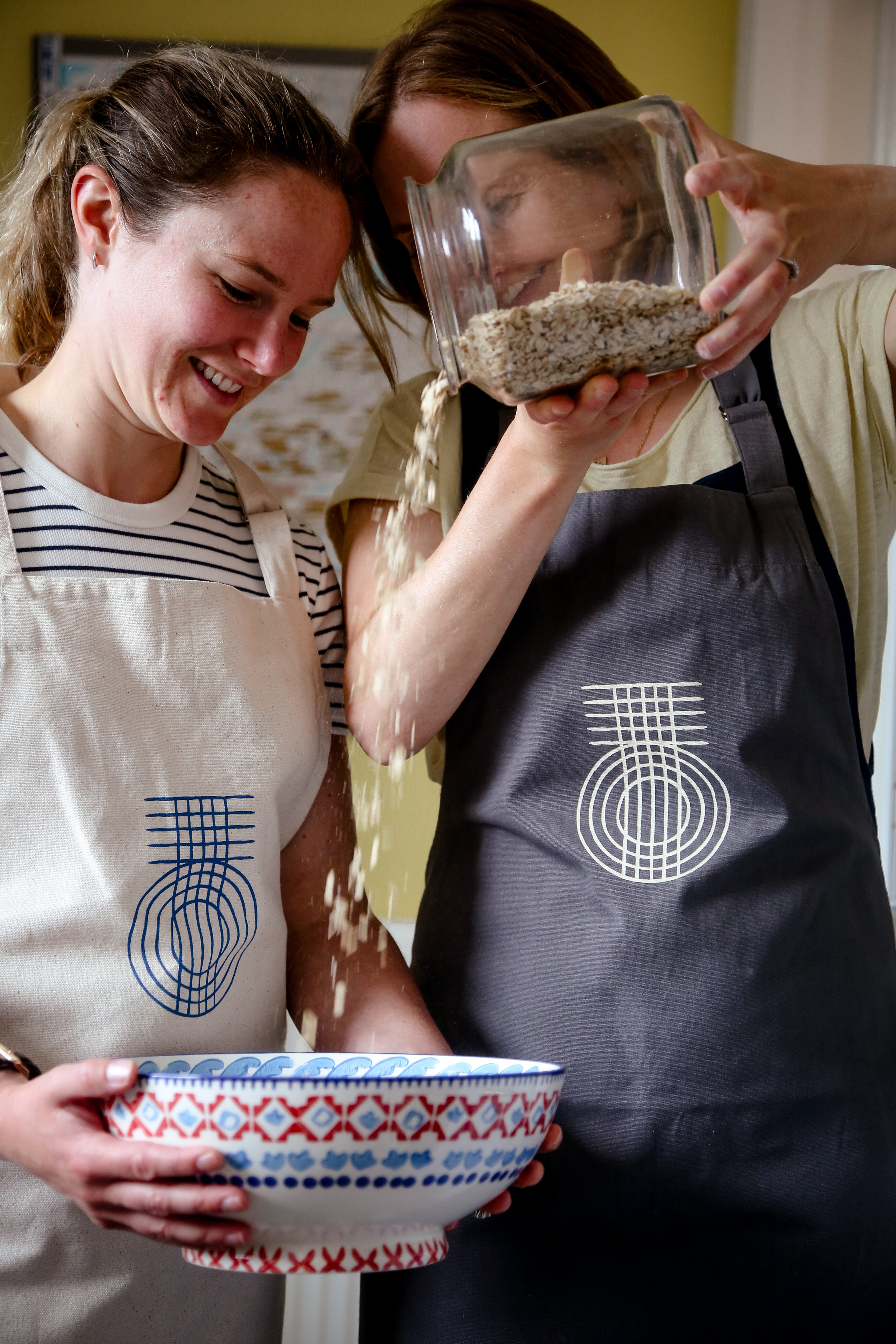 Two ladies having fun pouring ingredients into a bowl.  Both are wearing the True Origin Fairtrade aprons, one in beige and the other is grey.  Both have the True Origin weave logo on the front.