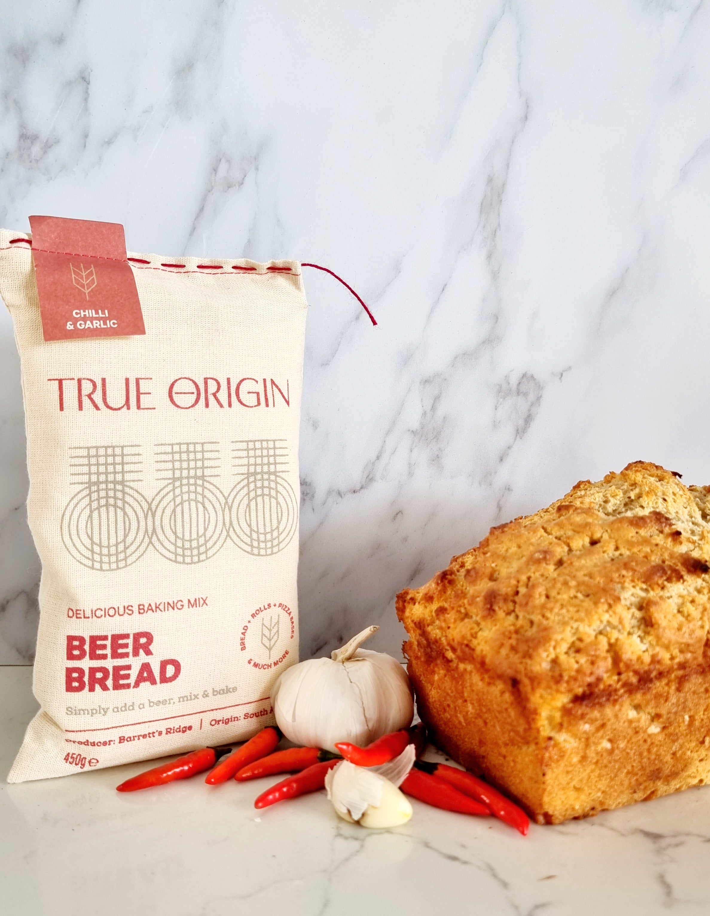 A bag of True Origin Garlic & Herb Beer Bread next to a freshly baked loaf of beer bread and a bulb of garlic and some chiilies.