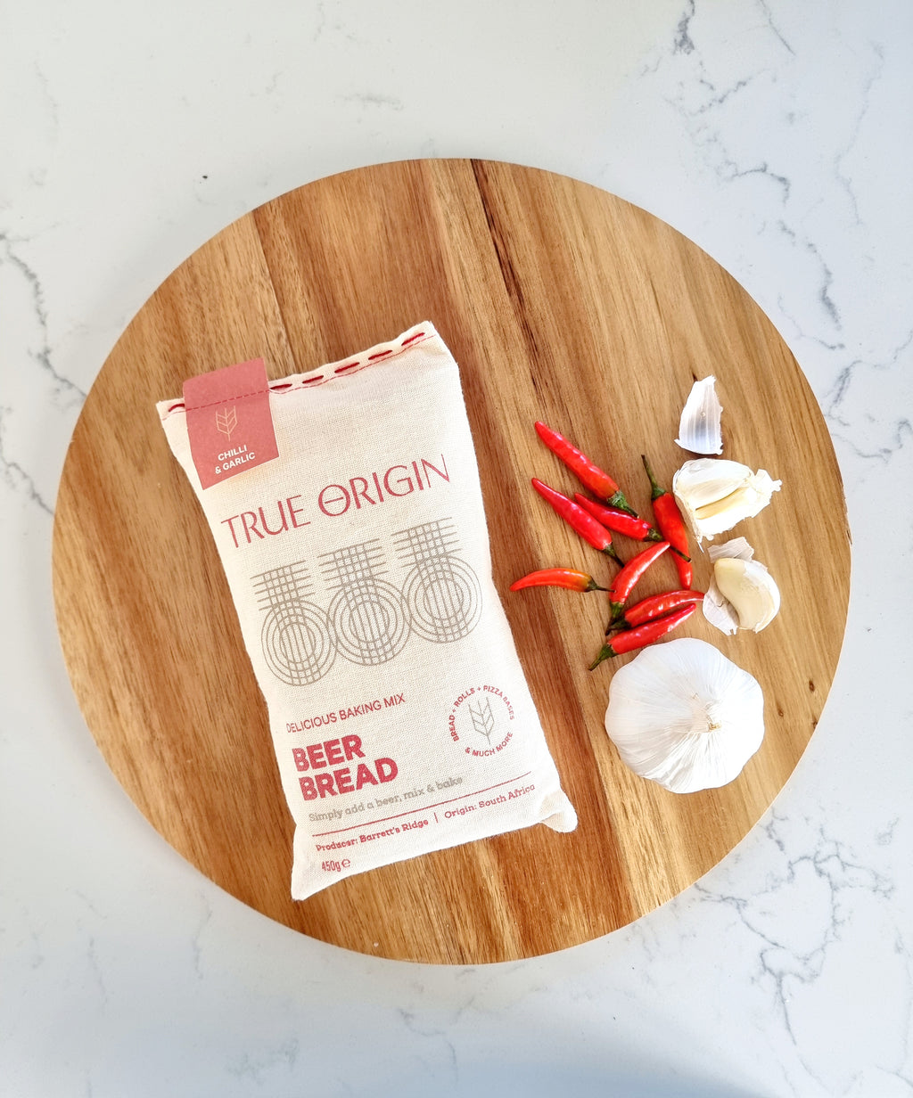 A bag of True Origin Chilli & Garlic Beer Bread on a circular wooden chopping board with a bulb of garlic and some fresh chillies.