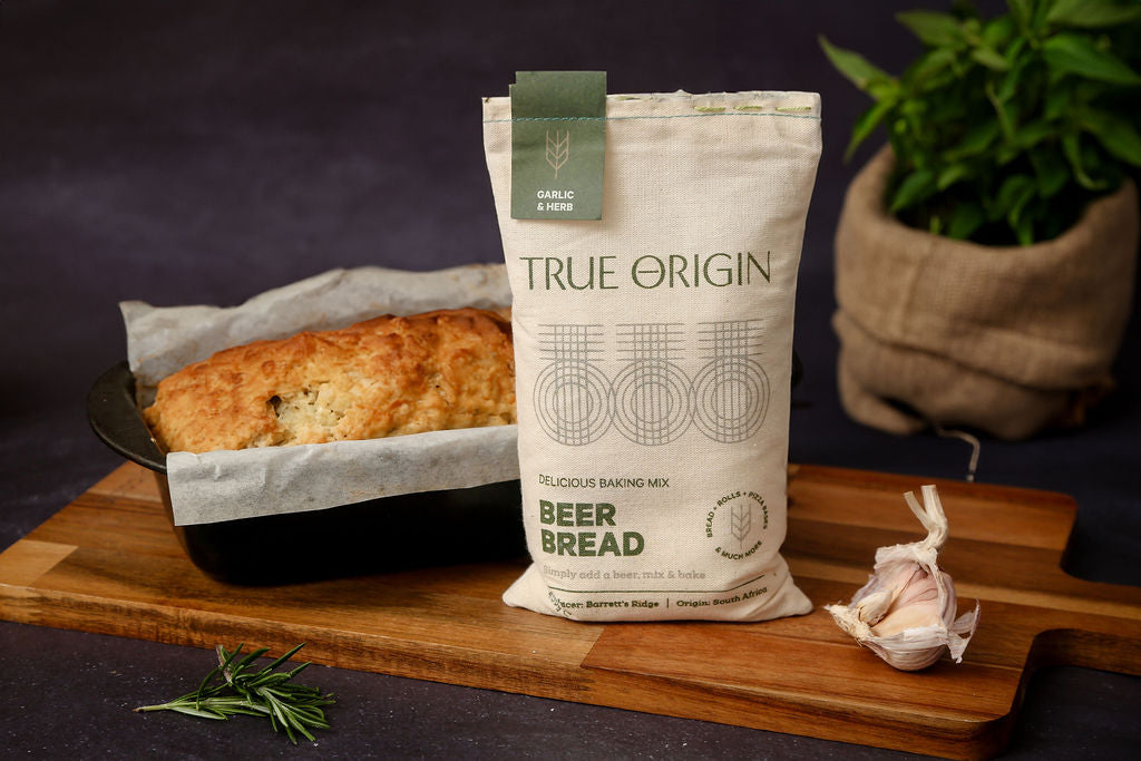 A bag of True Origin Garlic & Herb Beer Bread on a wooden chopping board with some fresh garlic and herbs.