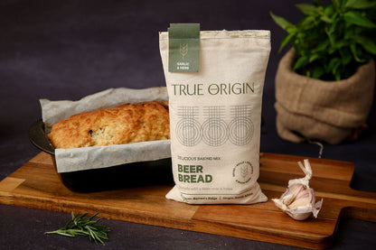 A bag of True Origin Garlic & Herb Beer Bread on a wooden chopping board with some fresh garlic and herbs.