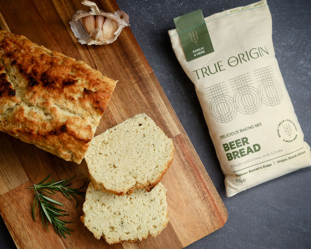 A bag of True Origin Garlic & Herb Beer Bread on a wooden chopping board next to a freshly baked loaf and slices with some fresh garlic and herbs.
