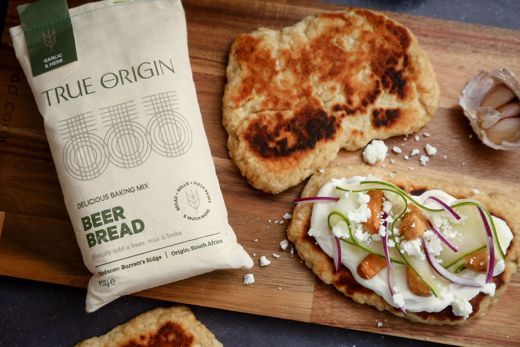 A bag of True Origin Garlic & Herb Beer Bread on a wooden chopping board with a flatbread made out of garlic and herb beer bread.
