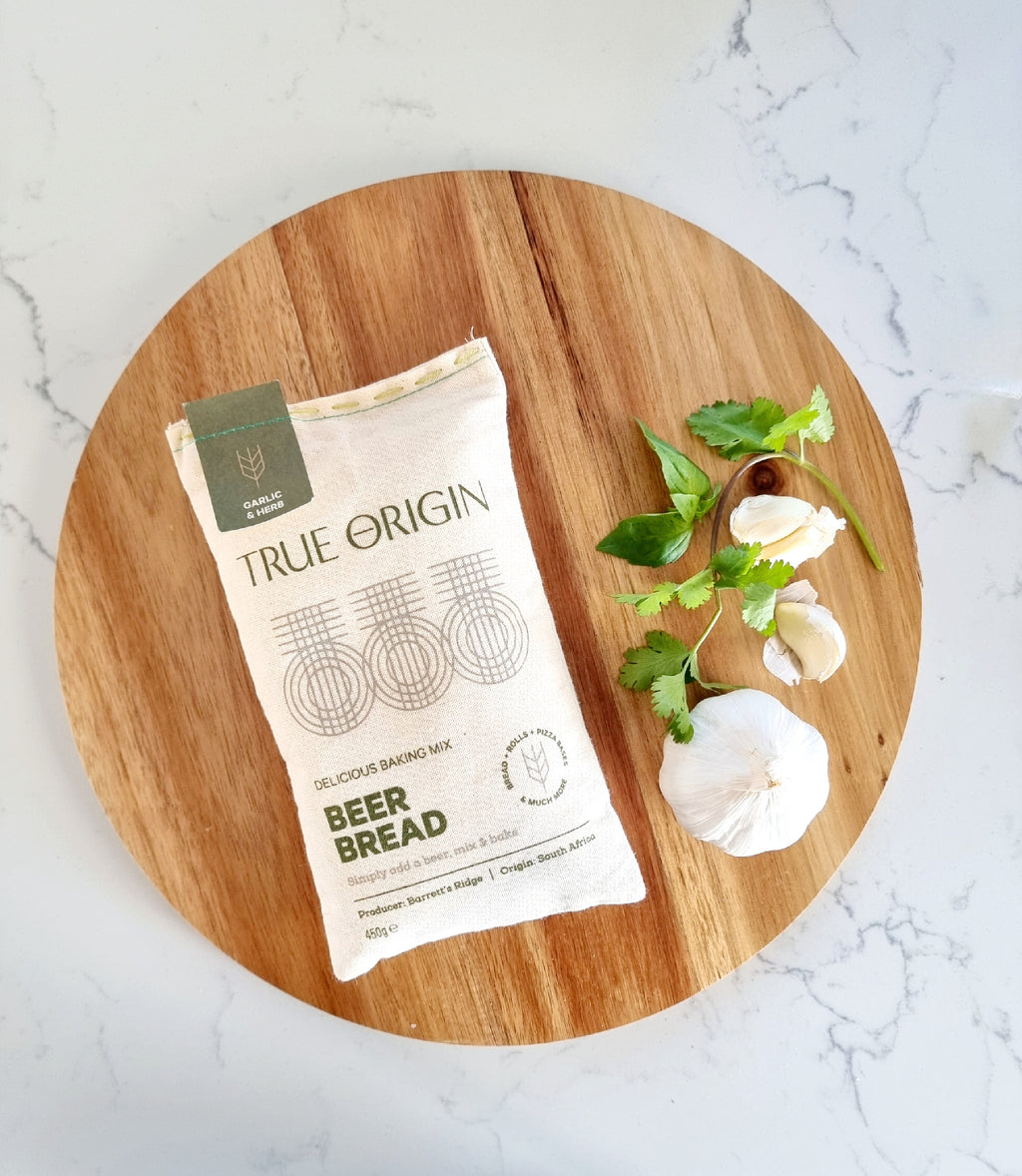 A bag of True Origin Garlic & Herb Beer Bread on a circular wooden chopping board with a bulb of garlic and some herbs.