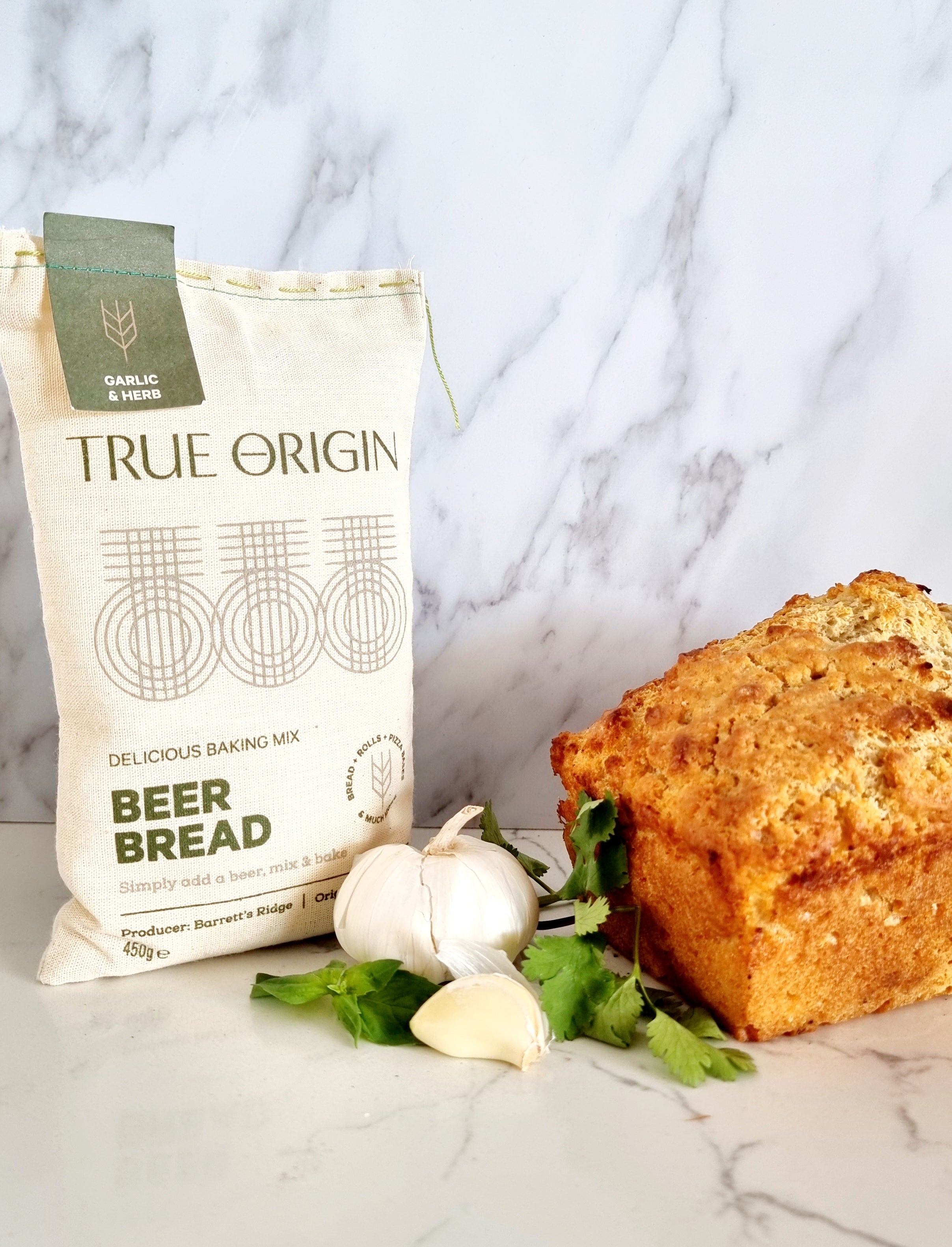 A bag of True Origin Garlic & Herb Beer Bread next to a freshly baked loaf of beer bread and a bulb of garlic and some herbs.