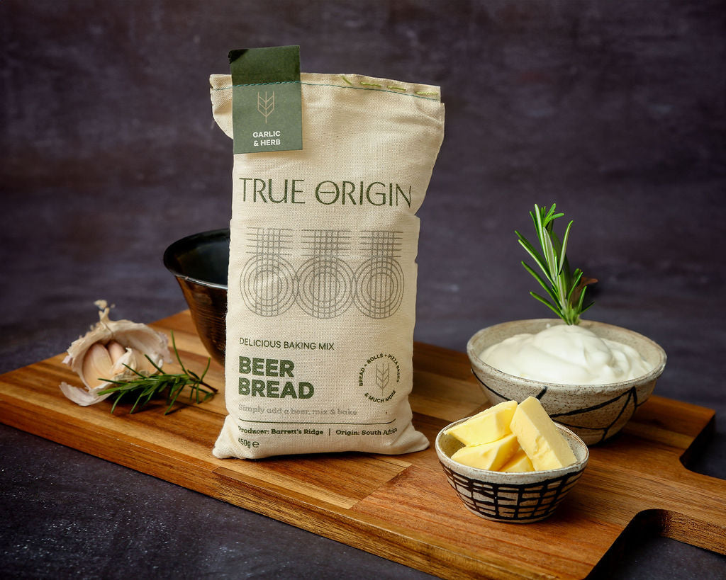 A bag of True Origin Garlic & Herb Beer Bread on a wooden chopping board with a bowl of butter and some fresh garlic and herbs.
