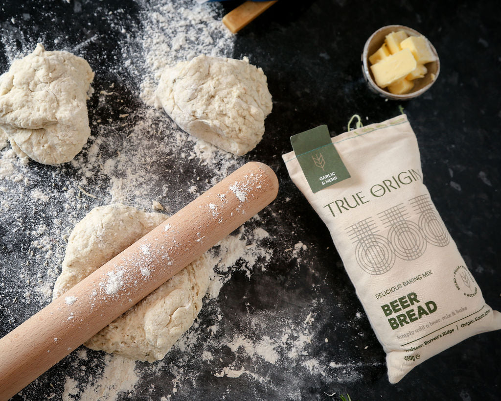 A bag of True Origin Garlic & Herb Beer Bread on a floured surface with a bowl of butter and balls of garlic and herbs beer bread dough.