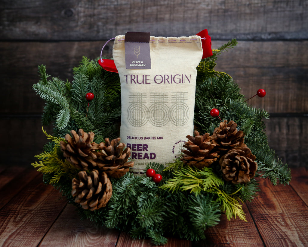 True Origin Olive & Rosemary beer bread mix package surrounded by festive greenery and pinecones on a wooden surface.