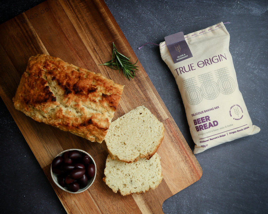 Loaf of olive and rosemary beer bread on a wooden cutting board with a package labelled 'True Origin olive and rosemary Beer Bread' in the background.