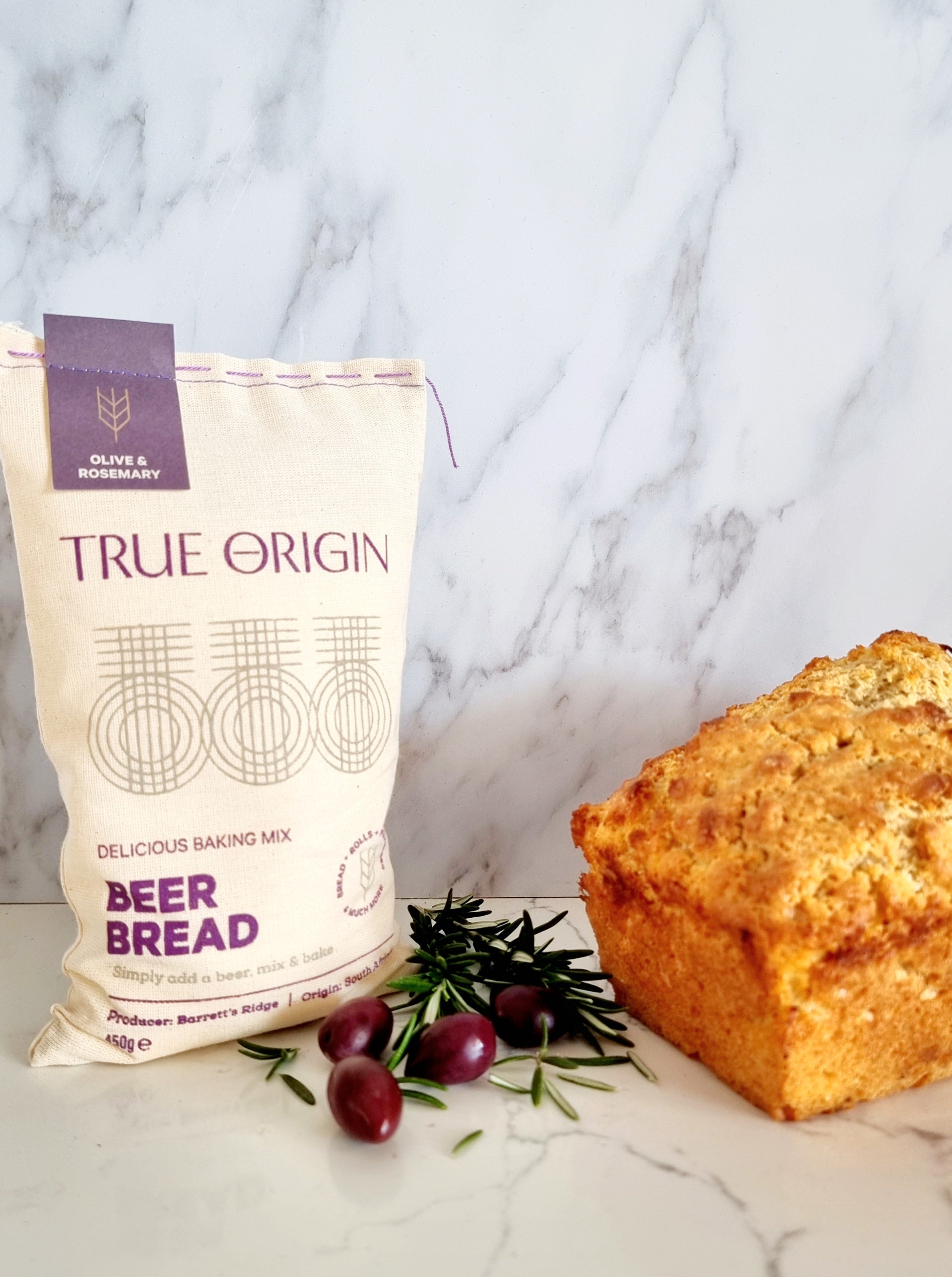 A bag of True Origin Olive & Rosemary Beer Bread next to a freshly baked loaf of beer bread and some fresh olives and rosemary.