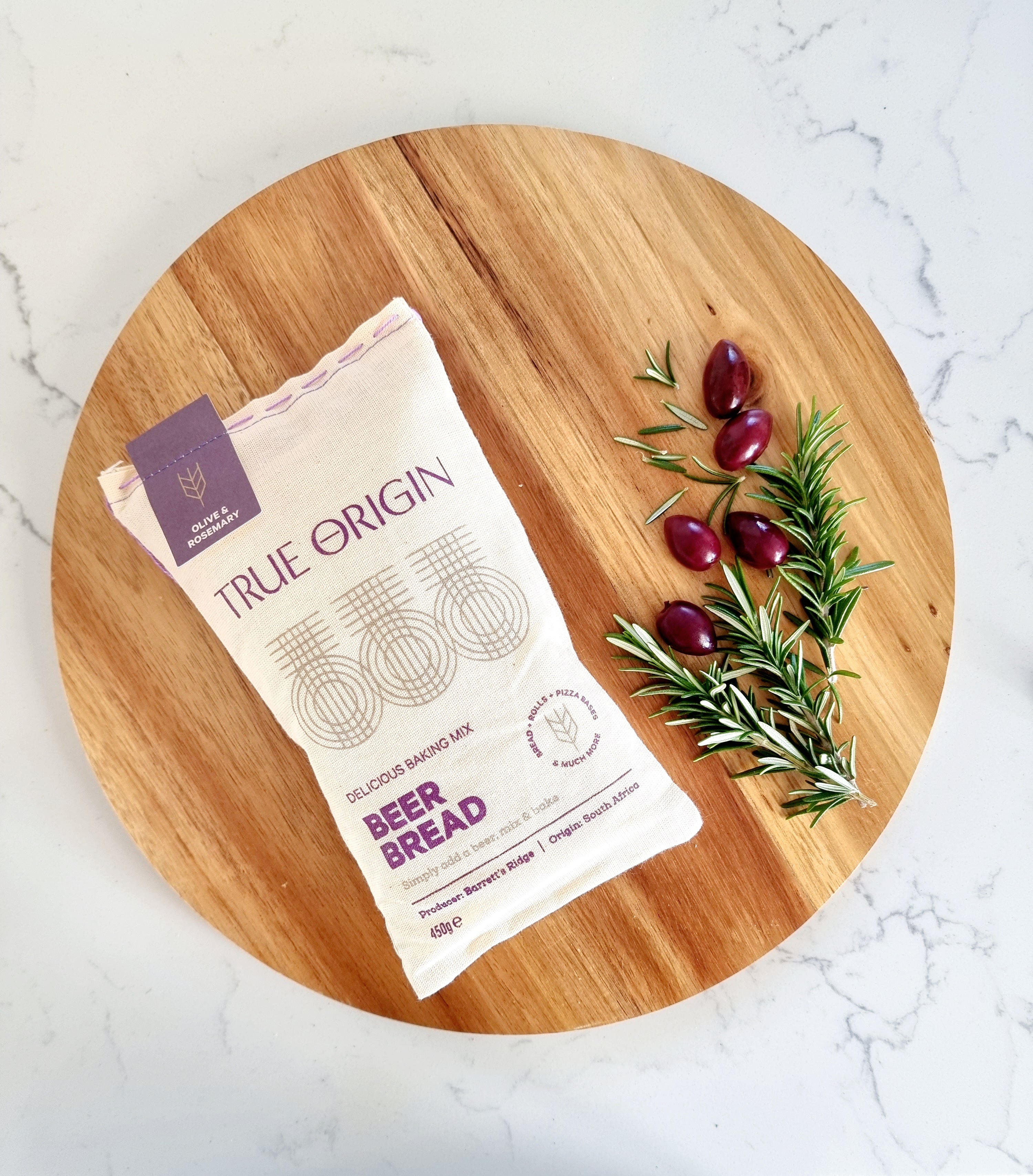 A bag of True Origin Olive & Rosemary Beer Bread on a circular wooden chopping board with some fresh olives and rosemary.