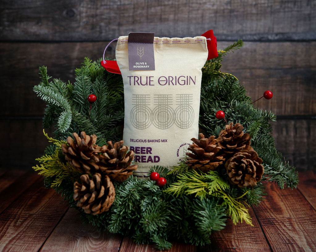 True Origin Olive & Rosemary Beer Bread mix package from South Africa surrounded by festive greenery and pinecones on a wooden surface.