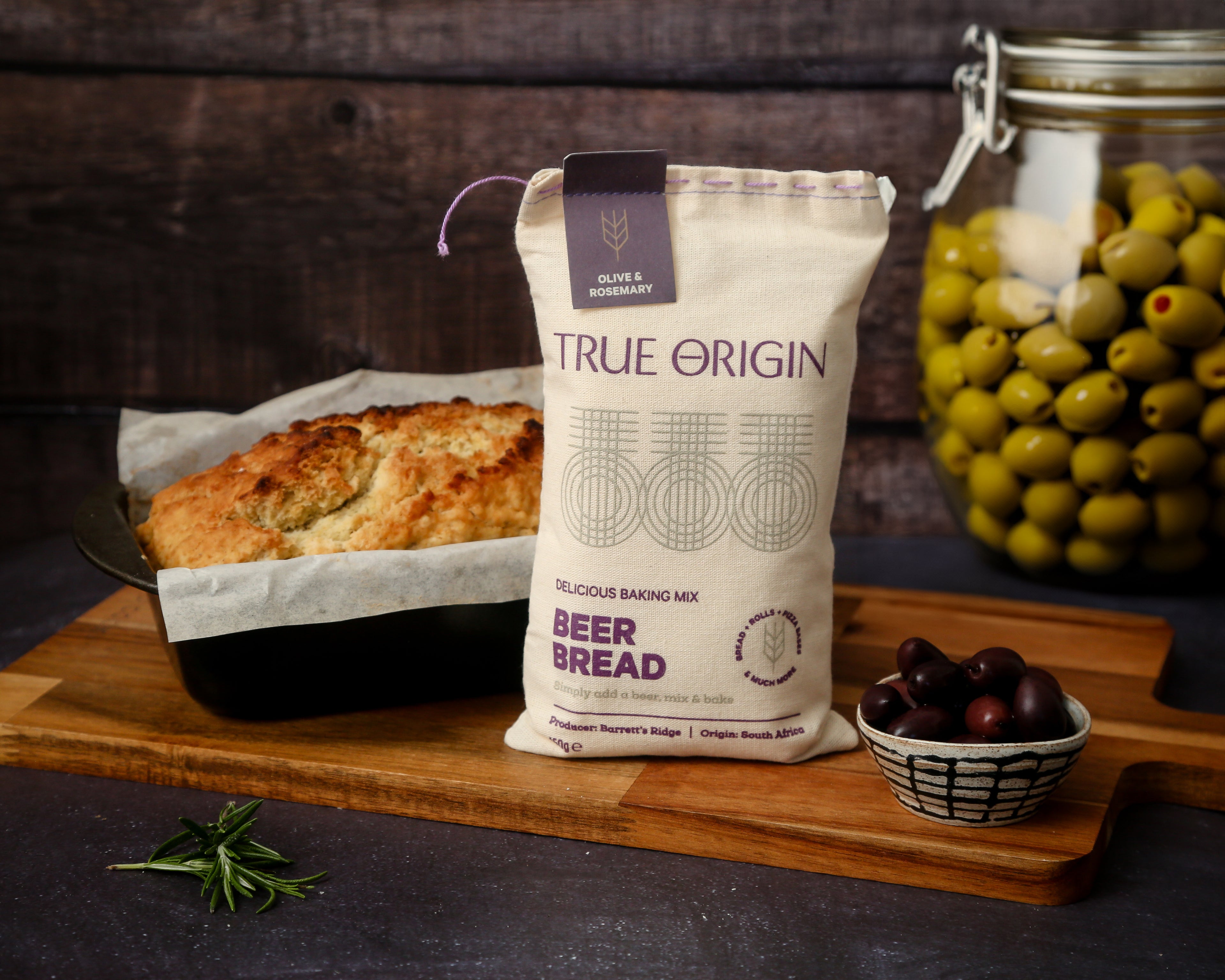 Bag of True Origin Olive & Rosemary beer bread baking mix on a wooden board with bread and olives in the background.