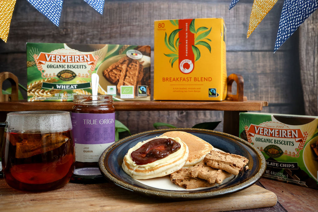 Breakfast setting with Vermeiren biscuits, Traidcraft tea, and a plate of pancakes with fair trade guava jam on a wooden table.