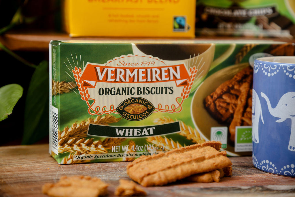 The bottom of a box of Traidcraft Fairtrade Tea, a packet of Vermeiren Organic Wheat Biscuits with a few loose biscuits and a blue mug with a white elephant design.