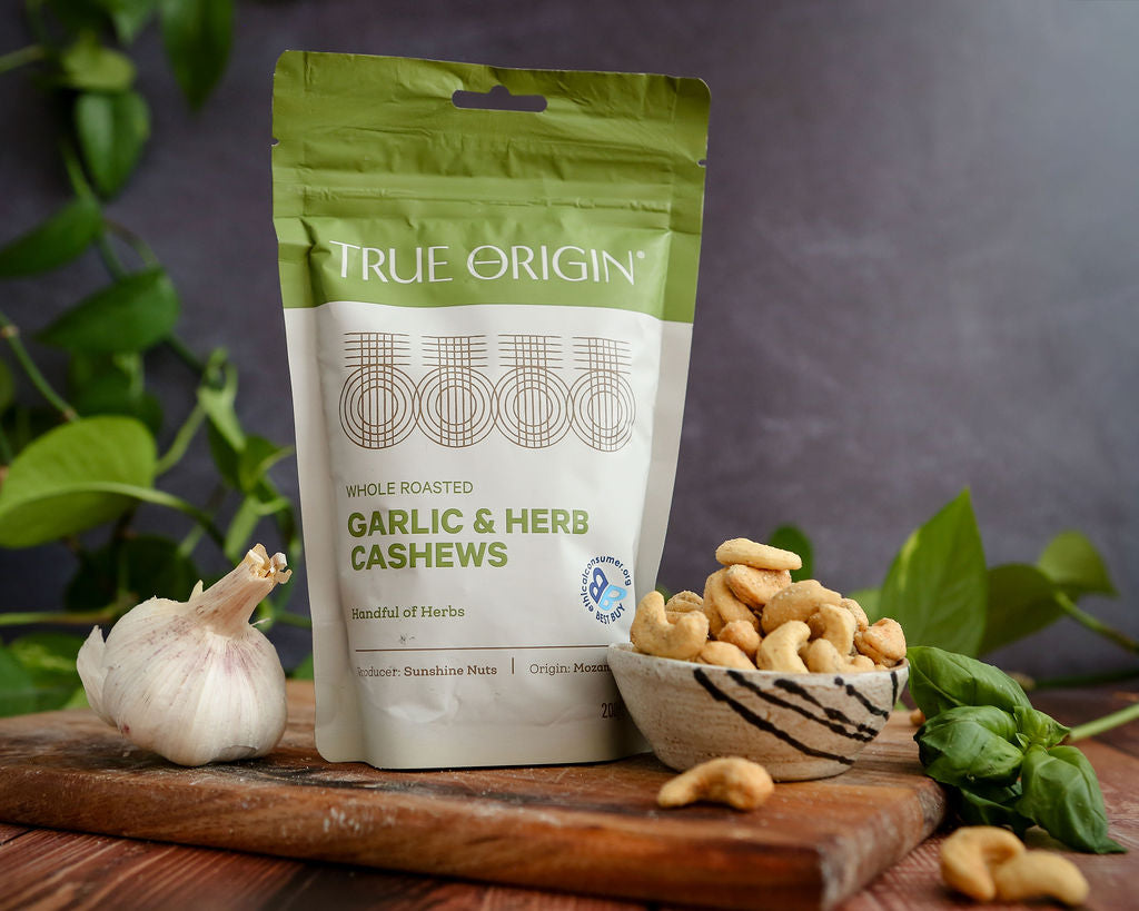 True Origin garlic and herb cashews packaging on a wooden board with cashews and garlic.