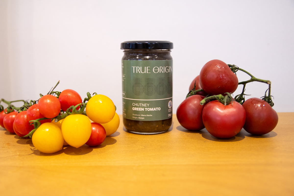 A jar of True Origin Chutney Green Tomato sits on a wooden table, flanked by fresh vine-ripe tomatoes in red, yellow, and dark red.