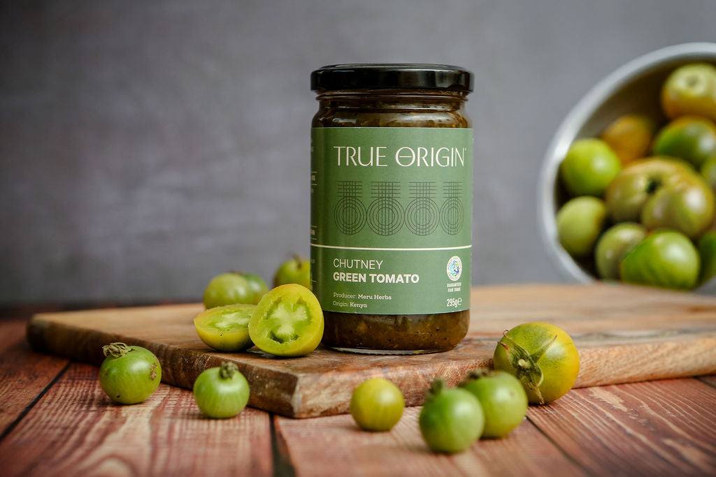 A jar of TRUE ORIGIN Chutney Green Tomato on a rustic wooden cutting board with fresh green tomatoes, set against a dark gray background with a bowl of tomatoes in the soft focus.