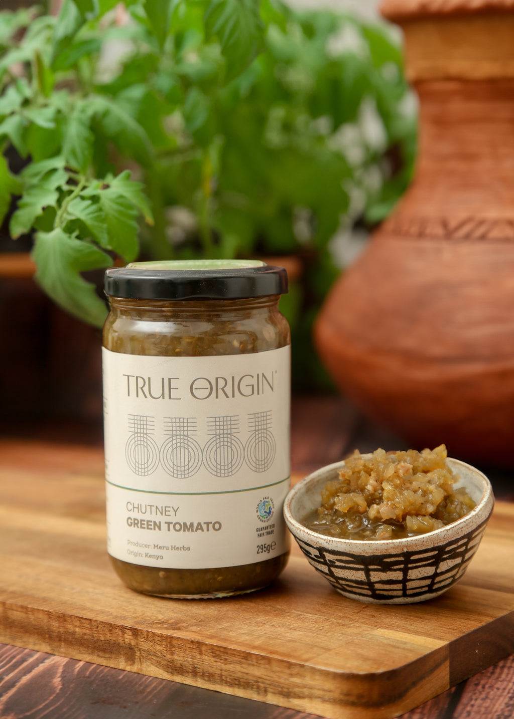 A jar of True Origin Green Tomato Chutney sitting on a wooden surface with a bowl of green tomato chutney and a bowl of fresh herbs.