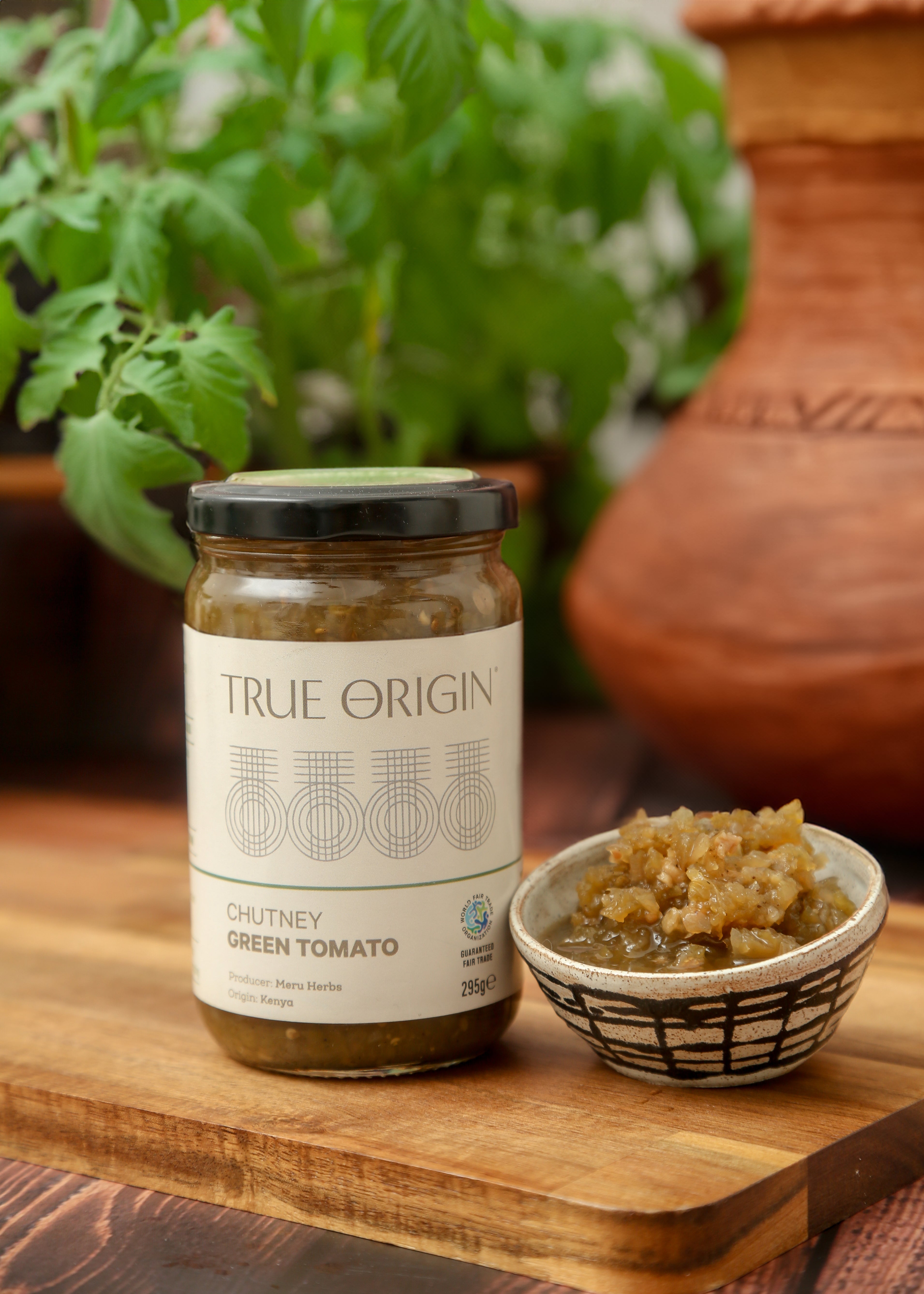 A jar of True Origin Green Tomato Chutney sitting on a wooden surface with a bowl of green tomato chutney and a bowl of fresh herbs.